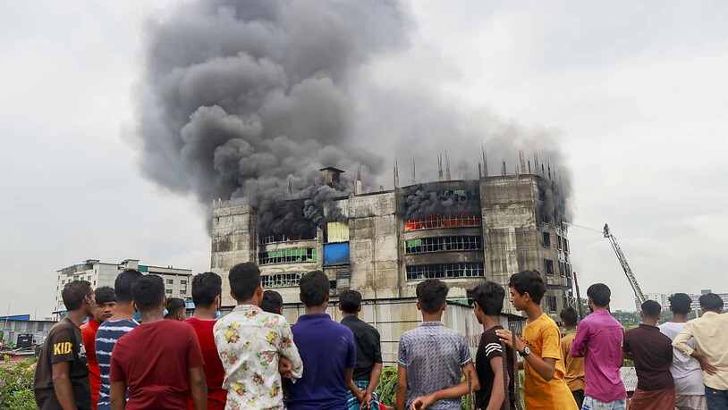 Menschen sehen zu, wie während des Brands in einer Fabrik Rauch von dem Gebäude aufsteigt. Bei einem Brand in einer Lebensmittel- und Getränkefabrik außerhalb der bangladeschischen Hauptstadt Dhaka sind mindestens 52 Menschen getötet worden. Foto: Suvra…